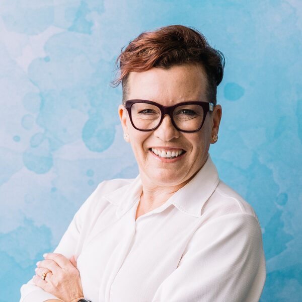 Female CEO smiling at the camera, wearing clear glasses with a shortred pixie haircut, white blouse and posing in front of a blue background
