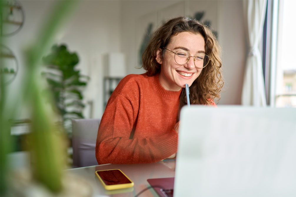 Happy young woman using laptop sitting at desk writing notes while watching webinar, studying online, looking at pc screen learning web classes or having virtual call meeting remote working from home.
