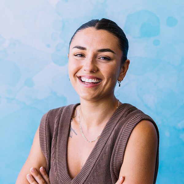 Marketing Specialist, Alycia Davies, Female Smiling at the camera in a brown knitted blouse and a blue backdrop.