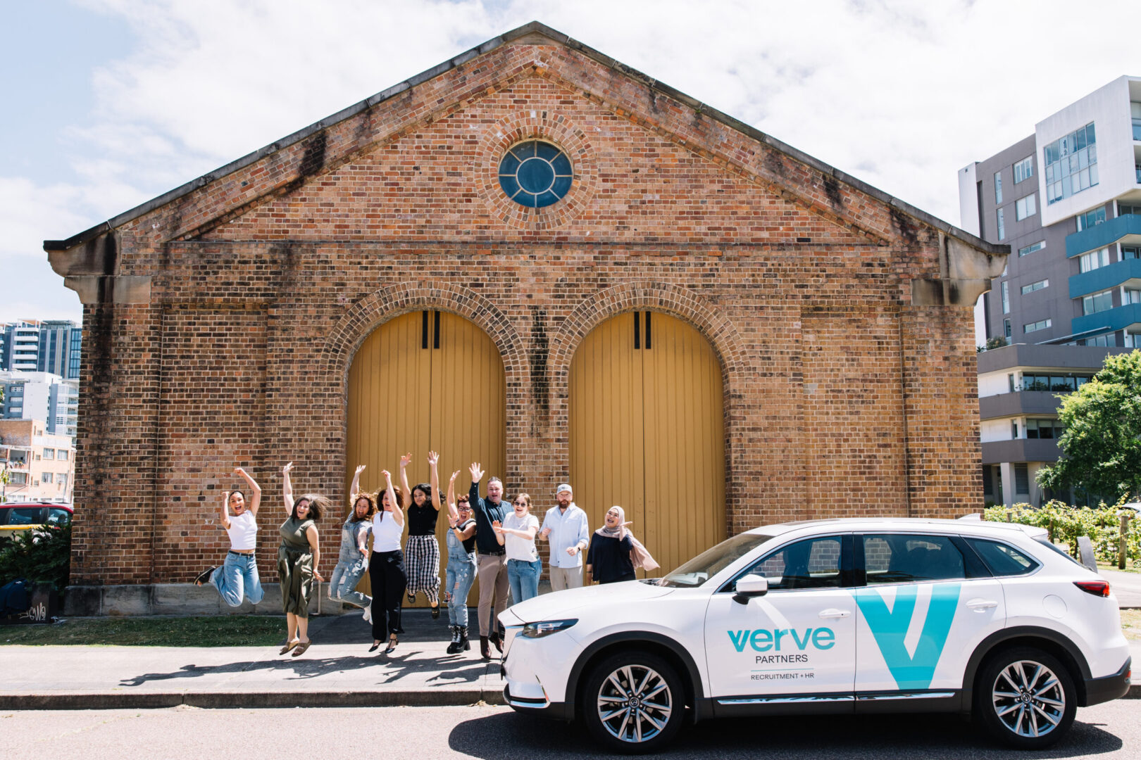 Newcastle Recruitment Career Opportunities At Verve Partners, Image shows Verve Team Members Jumping in the air for a posed group shot, with Verve Partners Company Vehicle in front of a historical building in the Newcastle CBD